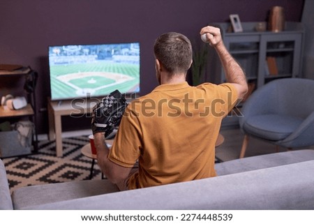 Back view at baseball fan watching match on TV at home and holding baseball ball, copy space