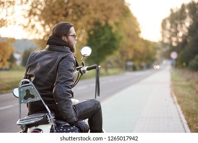 Back View Of Bearded Biker With Long Hair In Black Leather Jacket And Sunglasses Sitting On Modern Motorcycle On Blurred Background Of Straight Road Stretching To Horizon And Golden Autumn Trees.