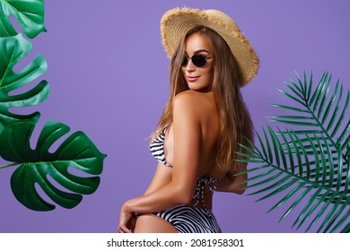 Back view of beautiful attractive young woman wears in bikini, straw hat and sunglasses who posing between tropical plants isolated on purple studio background  - Powered by Shutterstock