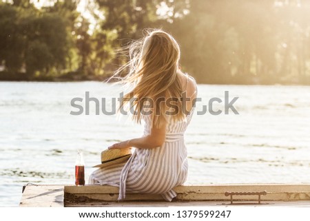 Back view of beautiful blonde girl in stylish stripped overalls sitting on wooden pier on summer sunset. Attractive young girl in stylish clothing posing with hair in motion on seaside. Hair in motion