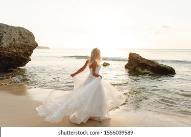 Back view of beautiful bride in white dress at sunset on ocean or sea coast on exotic island near water. Sun horizon. Sunny skyline. Wedding, honeymoon concept. Bride wearing beautiful wedding dress