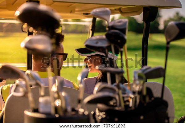 Back view of beautiful couple\
talking while driving a golf cart, golf clubs in the\
foreground