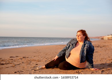 Back View Of Beautiful Overweight Woman Sitting On The Sandy Beach. Plus Size Girl Enjoy Warmth Sunset With Romantic Mood. Fat Model Dressed Jeans Jacket And Pink Knitted Sweater