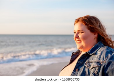 Back View Of Beautiful Overweight Woman Walking On The Sandy Beach. Plus Size Girl Enjoy Warmth Sunset With Romantic Mood. Fat Model Dressed Jeans Jacket And Pink Knitted Sweater