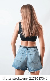 Back view of the beautiful young model in a casual black top and jeans shorts against  a grey background