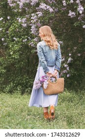 Back View Beautiful Young Stylish Blonde Girl In Denim Jacket And Dress Posing With Lilac Flowers In Straw Bag On Blossom Garden. Happy Romantic Attractive Hipster Woman Relax In Countryside Park