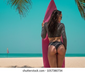 Back view of beautiful young woman in swimsuit and sunglasses posing with pink surfboard during sunny summer day on the tropical sandy beach over sea and sky background