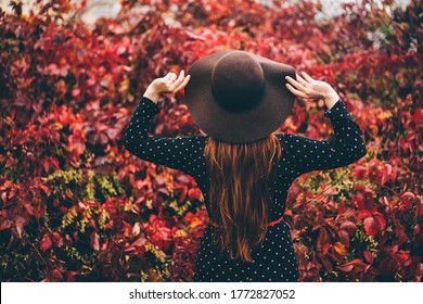 Back view of beautiful young woman in the hats standing at the autumn street. Trendy young red hair woman in fall in park. Concept of autumn mood.