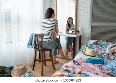 back view beauty asian woman doing makeup sitting at dressing table in bedroom. young girl looking in mirror and applying cosmetic with big brush. suitcase with cloth on floor packing for holiday - Powered by Shutterstock