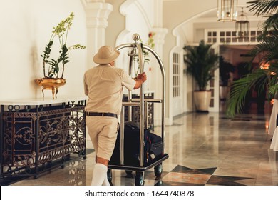 Back View Of Bellhop Pushing Luggage Cart Delivering Bags To Hotel Rooms.