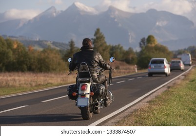 Back View Of Biker In Black Leather Jacket Riding Motorcycle Along Road On Blurred Background Of Beautiful Mountain Range With Snowy Peaks, Moving Vehicles On Bright Sunny Summer Evening.