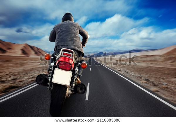 Back view of biker in\
motorcycle along road on blurred background of beautiful mountain\
range with snowy peaks, moving vehicles on bright sunny summer\
evening.