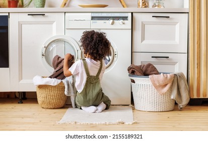 Back view of black boy with curly hair kneeling on floor and putting clothes into washing machine while doing laundry in kitchen at home