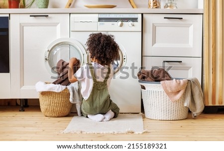 Back view of black boy with curly hair kneeling on floor and putting clothes into washing machine while doing laundry in kitchen at home