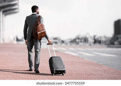 Back view of black businessman walking with rolling suitcase outside a modern building, ready for a business trip or travel. - Powered by Shutterstock