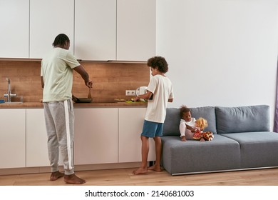 Back View Of Black Family Cooking Breakfast At Home. Father Frying Something On Pan While His Son Making Sandwich With Kiwi. Little Girl Sit And Play With Toys On Sofa. Fatherhood. Domestic Lifestyle