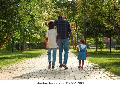 Back View Of Black Family Walking On Pavement Road In Green Park. Father Hugging Son And Holding Hand Of Daughter. Family Relationship And Spend Time Together. Fatherhood And Parenting. Warm Sunny Day