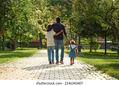 Back view of black family walking on pavement road in green sunny park. Father hug with son and hold hand of little daughter. Family relationship and spending time together. Fatherhood and parenting