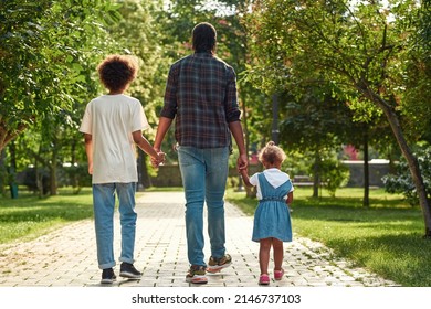 Back View Of Black Family Walking On Pavement Road In Green Park. Father Hold Hands Of Son And Little Daughter. Family Relationship And Spend Time Together. Fatherhood And Parenting. Warm Summer Day