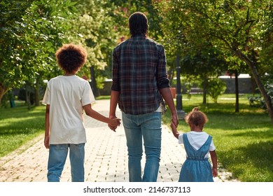 Back View Of Black Family Walking On Pavement Road In Green Park. Father Hold Hands Of Son And Little Daughter. Family Relationship And Spend Time Together. Fatherhood And Parenting. Sunny Summer Day