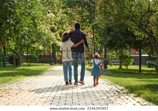 Back view of black family walking on pavement\
road in green sunny park. Father hug with son and hold hand of\
little daughter. Family relationship and spending time together.\
Fatherhood and parenting
