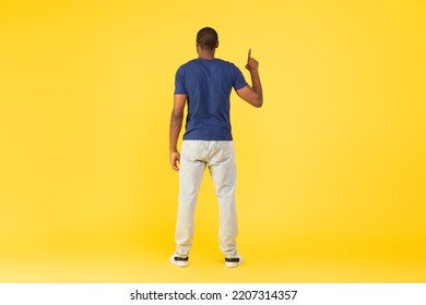 Back View Of Black Guy Pointing Finger Upward Standing In Studio, Pretending Pushing Invisible Button Or Touching Touchscreen Standing On Yellow Background. Look Up Concept - Powered by Shutterstock