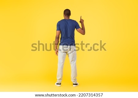 Back View Of Black Guy Pointing Finger Upward Standing In Studio, Pretending Pushing Invisible Button Or Touching Touchscreen Standing On Yellow Background. Look Up Concept