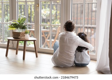 Back view of black haired young mom and little daughter girl sitting on floor at home, hugging with love, care, looking at panoramic window, cozy balcony. Motherhood, real estate concept - Powered by Shutterstock