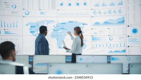 Back View Of Black Male Data Scientist Talking To Caucasian Female Supervisor In Front Of Digital Screen With World Map In Monitoring Office. Employees Working Behind Computers in Distribution Company