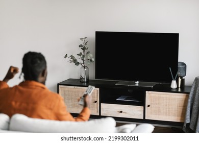Back View Of Black Man Watching Sports Game On TV, Celebrating Victory And Shaking Fists, Sitting On Sofa In Living Room. Television Set With Empty Screen. Mockup