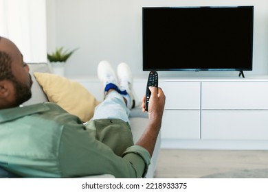 Back View Of Black Man Watching TV Pointing Remote Control At Empty Plasma Screen, Switching Channels, Resting On Sofa At Home. Guy Enjoying Movie Or Sport Program, Using Television Set. Mockup