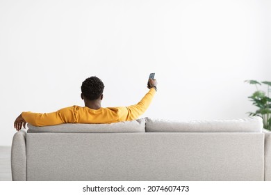 Back view of black man in yellow sitting on couch with remote from TV, pointing towards empty white wall, african american guy turning on air conditioner, home interior, copy space