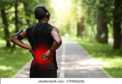 Back view of black sportsman touching sore zone on his back, having back pain during jogging by park, empty space