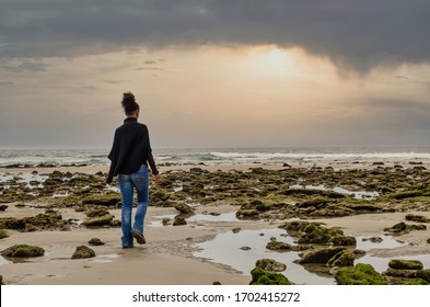 Back View Of A Black Woman Walking On Zahora Beach In Cadiz Between Moss Covered Rocks.  Selective Focus On The Rocks.  Long Exposure. Beautiful Sunset.