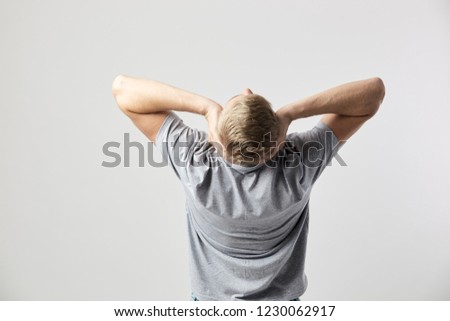 Back view of blond guy dressed in a white t-shirt that holds his hands on his head on the white background in the studio
