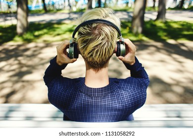 Back View Of Blond Man In Dark Clothes Sitting On White Sunlit Park Bench Putting Grey Headphones On Head On Blurred Background