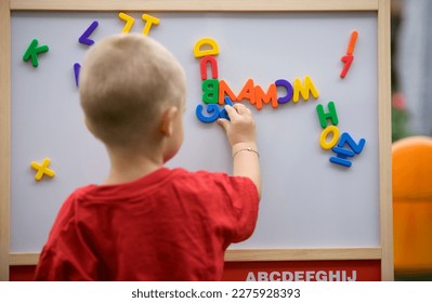 Back view of a blond toddler boy playing with colorful letters on a magnetic board trying to make a word  - Shutterstock ID 2275928393
