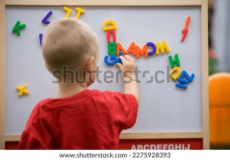 Back view of a blond toddler boy playing with colorful letters on a magnetic board trying to make a word 