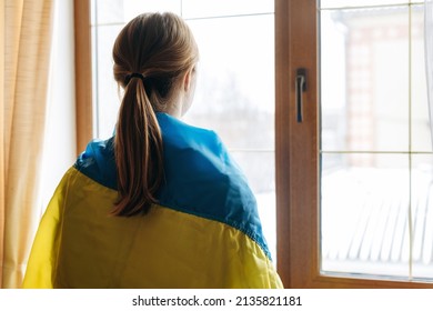 Back view of the blonde woman wrapped in Ukraine flag looking out of the window. Support for Ukraine concept 