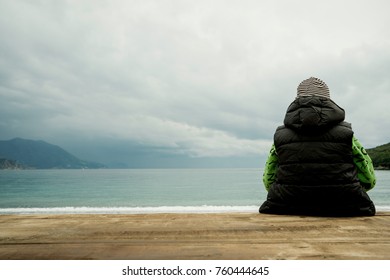 Back view of a boy sitting on a wooden board and looking at the sea