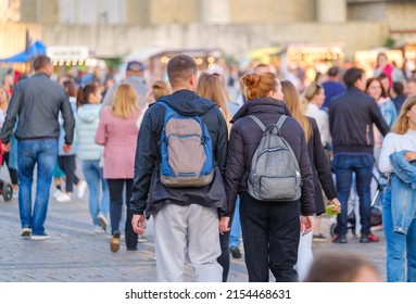Back view of boyfriend and girlfriend in outerwear with backpack looking away while strolling in crowded park during public event