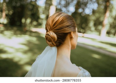 Back view of a bride with elegant blonde bun hairstyle and a wedding veil, walking outdoors in a sunlit green park or forest. Close-up