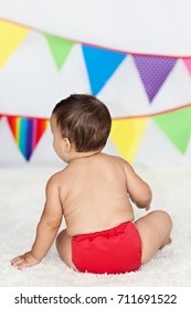 A Back View Of A Brown Haired Baby Boy Wearing A Red Cloth Diaper / Nappy Sitting On A White Carpet With A Brightly Colored Flag Bunting In The Background.