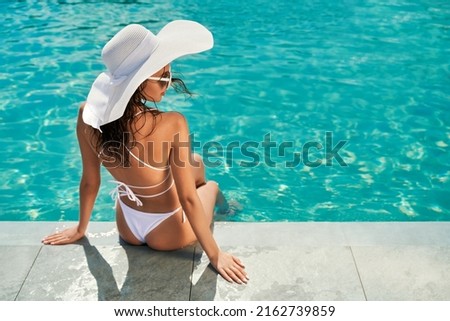 Back view of brunette girl wearing white swimsuit, hat and sunglasses, sunbathing. Slim seductive female sitting near swimming pool, relaxing, looking aside. Concept of vacation.