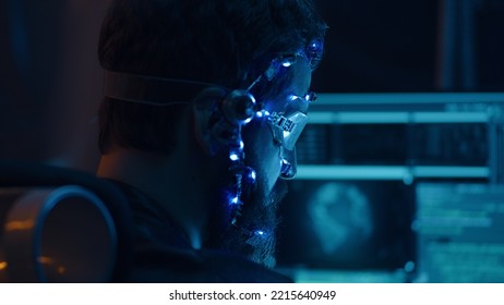 Back view of a brunette guy wearing futuristic headset with LED lights attached programs and develops code using multiple computer screen. Cyperpunk style. Sci-fi background. Neon lights.