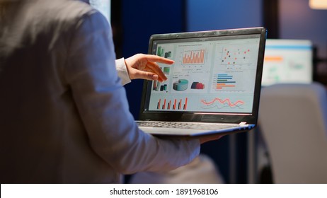 Back view of business woman analysing financial statistics holding laptop standing in start-up office late at night overworking. Busy focused employee using modern technology network wireless overtime