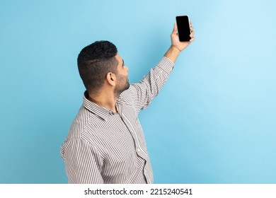 Back View Of Businessman Talking Video Call, Taking Selfie On Mobile Phone, Holding Smartphone Up With Mockup Blank Display, Wearing Striped Shirt. Indoor Studio Shot Isolated On Blue Background.