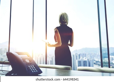 Back view of a businesswoman with digital tablet in hands is watching in office window on a Hong Kong city view and thinking about future her company. Female manager is using touch pad during work day