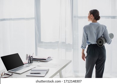 back view of businesswoman holding yoga mat at workspace with laptop and notepad - Powered by Shutterstock