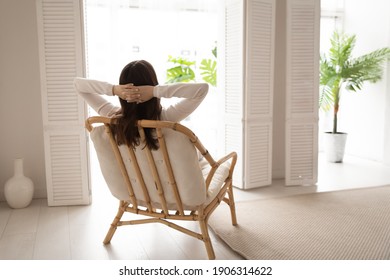 Back view of calm young woman sit rest in cozy modern chair in living room stretch breathe fresh air. Happy female renter relax in armchair at home look in window think or visualize enjoy weekend.